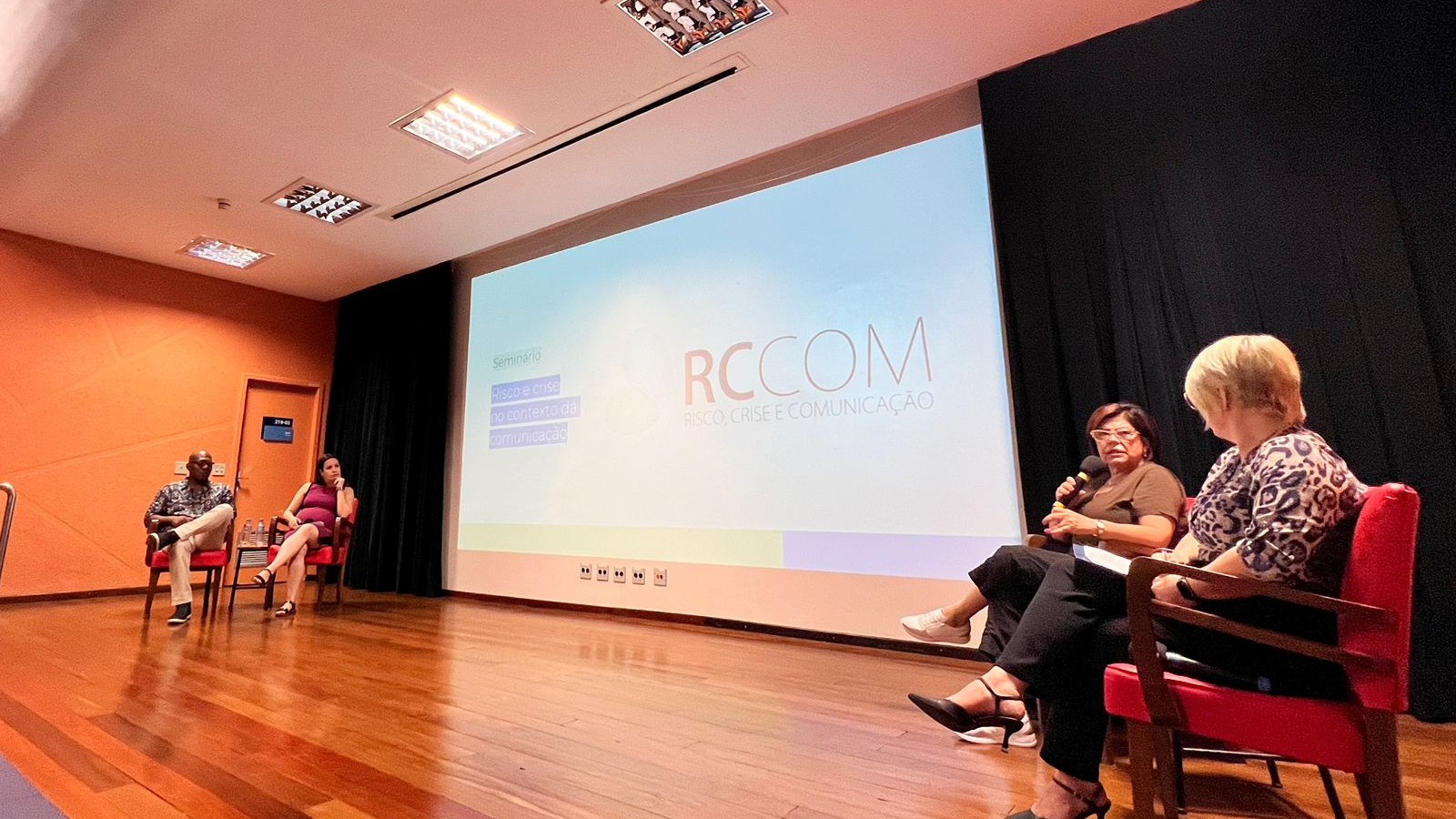 expositores convidados sentados em um palco em que a secretária Tânia Moreira segura o microfone