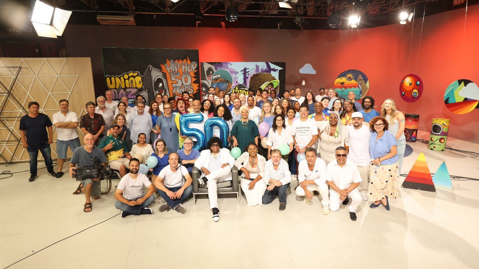 No estúdio de televisão, um grande grupo de pessoas posa para a fotos, no meio os balões com os números 5 e 0.