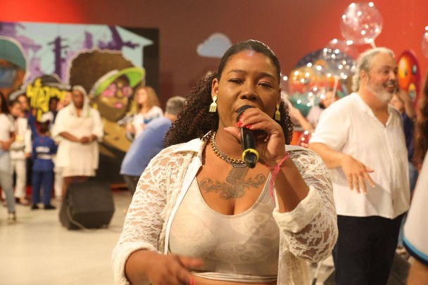 A cantora vestida de branco segura o microfone e atrás muitas pessoas confraternizam