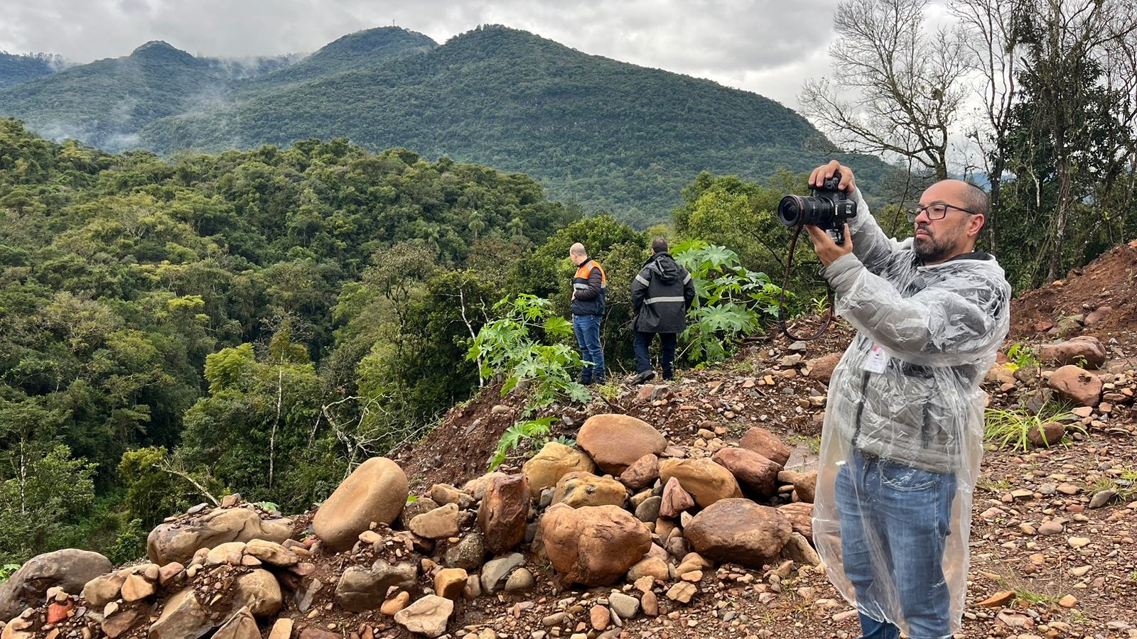 A foto mostra o repórter cinematográfico Luis André Pinto, um homem de barba e óculos, de pé no primeiro plano à direita. Ele veste uma capa de chuva plástica transparente e calça jeans, e está segurando uma câmera profissional com as duas mãos, apontando-a para longe. Ele está em uma área de terreno irregular, com muitas pedras e terra. No fundo, há uma vasta paisagem montanhosa coberta por densa vegetação verde, sob um céu nublado. Duas pessoas de costas são visíveis ao longe, no meio da vegetação. Uma delas veste o colete com faixa laranja da Defesa Civil. A cena retrata um trabalho de campo na região de Caxias do Sul.