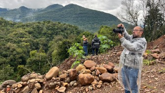A foto mostra o repórter cinematográfico Luis André Pinto, um homem de barba e óculos, de pé no primeiro plano à direita. Ele veste uma capa de chuva plástica transparente e calça jeans, e está segurando uma câmera profissional com as duas mãos, apontando-a para longe. Ele está em uma área de terreno irregular, com muitas pedras e terra. No fundo, há uma vasta paisagem montanhosa coberta por densa vegetação verde, sob um céu nublado. Duas pessoas de costas são visíveis ao longe, no meio da vegetação. Uma delas veste o colete com faixa laranja da Defesa Civil. A cena retrata um trabalho de campo na região de Caxias do Sul.