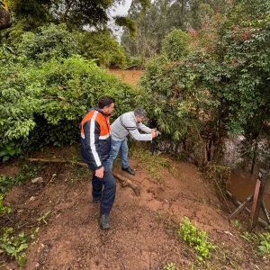 A foto mostra dois homens em uma área rural com terreno enlameado e vegetação densa. À esquerda, um homem com colete laranja e azul da Defesa Civil e calça escura está de costas, olhando para a frente. À direita e ligeiramente à frente dele, o repórter fotográfico Igor de Almeida de camisa cinza listrada e jeans está agachado, apontando uma câmera fotográfica para um rio com águas barrentas, visível no canto inferior direito. O rio está cercado por folhagens e árvores de bambu. O céu está nublado, indicando um dia chuvoso ou pós-chuva, e a cena sugere a documentação de uma área afetada por eventos meteorológicos.