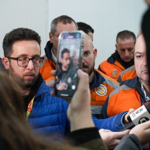 A foto mostra um close de uma cena de reportagem onde um smartphone é levantado no primeiro plano com a imagem desfocada do entrevistado em close. Há vários microfones sendo segurados em direção ao entrevistado. No centro da imagem, em destaque, o repórter Carlos Ismael Moreira. Ele é um homem barbudo de óculos e jaqueta azul. Ele é o foco principal e está olhando para a frente, parecendo estar em uma coletiva de imprensa ou entrevista. Ao fundo, outros homens de coletes laranja e azul da Defesa Civil. A cena transmite a intensidade de uma cobertura jornalística.