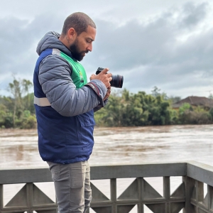 A foto mostra o repórter Brayan Martins, um homem com barba e cabeça raspada, de perfil, em pé em um deck de madeira em frente a um rio de águas barrentas. Ele veste uma jaqueta com cores cinza, verde e azul, calças cinzas e botas de borracha pretas com detalhes amarelos. Brayan Martins está concentrado em operar uma câmera fotográfica profissional que segura com ambas as mãos, direcionando a lente para algo fora do campo de visão à sua frente. Ao fundo, a margem oposta do rio é coberta por vegetação densa e árvores sob um céu nublado e uniforme. A cena sugere a cobertura jornalística de uma área possivelmente afetada por enchentes ou outros eventos naturais.