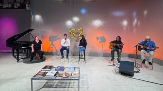 Foto mostra o est&uacute;dio do Programa Esta&ccedil;&atilde;o Cultura Clube Especial 52 anos, com entrevistados e apresentadores, al&eacute;m de dois m&uacute;sicos. H&aacute; um piano e uma mesa com livros. 